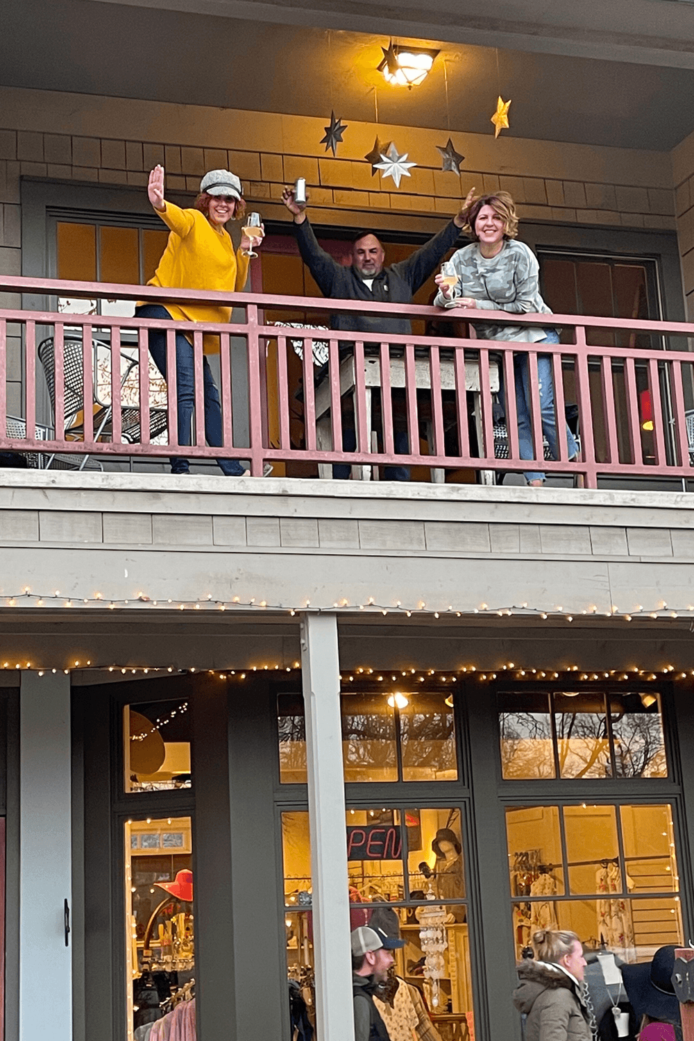 Three people happily wave from a balcony adorned with stars, while guests stroll below.